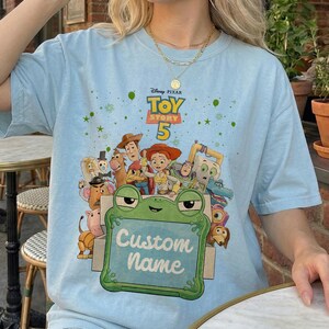 Puede incluir: Camiseta azul claro con un gr&aacute;fico de personajes de la pel&iacute;cula de Disney Pixar Toy Story 5. El dise&ntilde;o incluye a Woody, Buzz Lightyear y otros personajes. Un letrero verde con forma de rana dice "Custom Name". La camiseta tiene el logo de Toy Story 5.