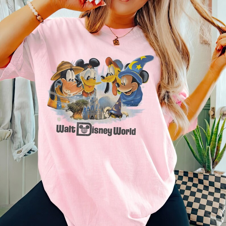 Pu&ograve; includere: T-shirt rosa chiaro con una grafica dei personaggi Disney: Pippo, Paperino, Pluto e Topolino con un cappello da mago. Il testo "Walt Disney World" &egrave; stampato sotto in nero.