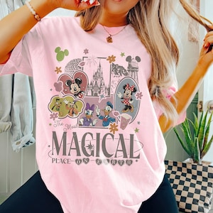 May include: Light pink t-shirt featuring a graphic design with Mickey and Minnie Mouse, Donald Duck, Daisy Duck, and Goofy. The design includes the text "The Most Magical Place on Earth" in a decorative font.