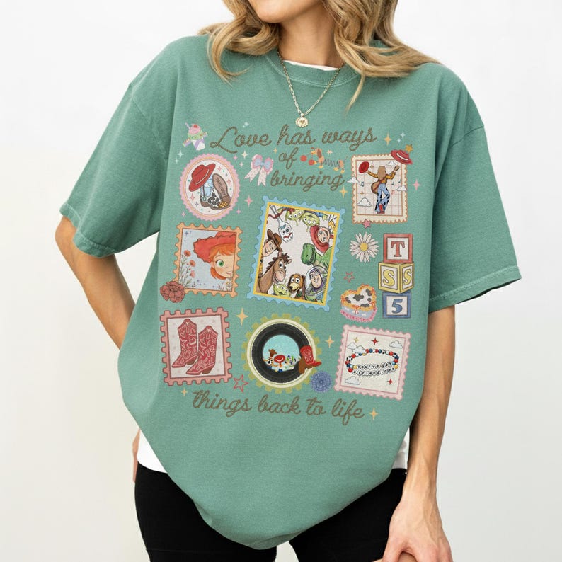 May include: A sage green short-sleeve t-shirt featuring a graphic print with the text "Love has ways of bringing things back to life." The design includes various postage stamp-style frames containing illustrations of cowboy boots, characters, and toys.