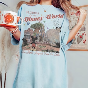Puede incluir: Camiseta azul claro con un gr&aacute;fico de Florida Disney World. El dise&ntilde;o presenta a Mickey Mouse, la Torre Hollywood y las palabras "The Most Magical Place On Earth". Una taza con un dise&ntilde;o de sol se sostiene en la mano.