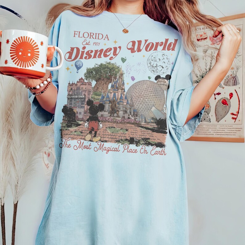 Puede incluir: Camiseta azul claro con un gr&aacute;fico de Florida Disney World. El dise&ntilde;o presenta a Mickey Mouse, la Torre Hollywood y las palabras "The Most Magical Place On Earth". Una taza con un dise&ntilde;o de sol se sostiene en la mano.