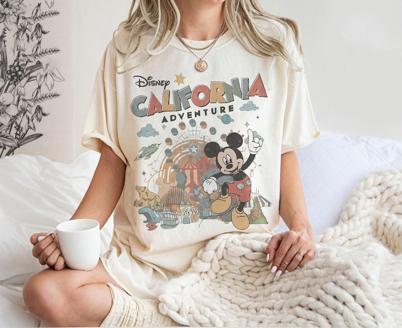 Puede incluir: Camiseta color crema con un gr&aacute;fico de Mickey Mouse y las palabras "California Adventure" en letras coloridas. El dise&ntilde;o incluye im&aacute;genes de una noria, un cohete y otros elementos de parque de atracciones. La camiseta es de corte holgado.