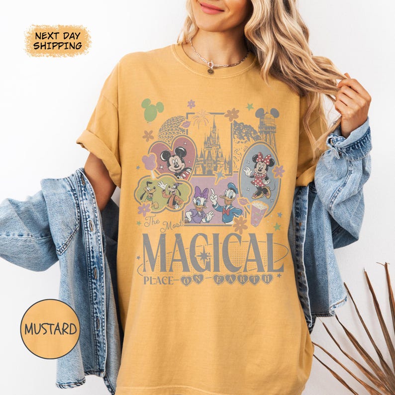 May include: Mustard-colored t-shirt featuring a colorful graphic of Disney characters, including Mickey and Minnie Mouse, Donald and Daisy Duck, and Goofy. The graphic includes the text "The Most Magical Place on Earth."