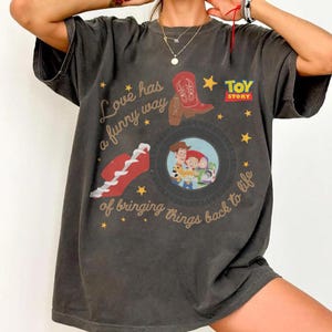 May include: A charcoal gray short-sleeve t-shirt featuring a Toy Story graphic with Woody, Jessie, and Buzz Lightyear inside a tire. The design includes a red cowboy boot, a red sneaker, yellow stars, and the text Love has a funny way of bringing things back to life.