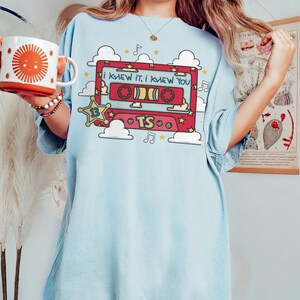 May include: A light blue t-shirt featuring a graphic of a red cassette tape with the text "I KNEW IT, I KNEW YOU" and "TS" with "13" inside a star. The design includes white clouds, yellow stars, and musical notes. A white mug with an orange sun is visible.