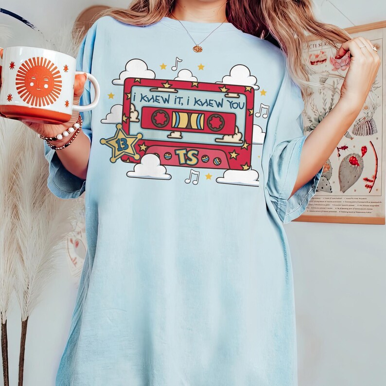 May include: A light blue t-shirt featuring a graphic of a red cassette tape with the text "I KNEW IT, I KNEW YOU" and "TS" with "13" inside a star. The design includes white clouds, yellow stars, and musical notes. A white mug with an orange sun is visible.