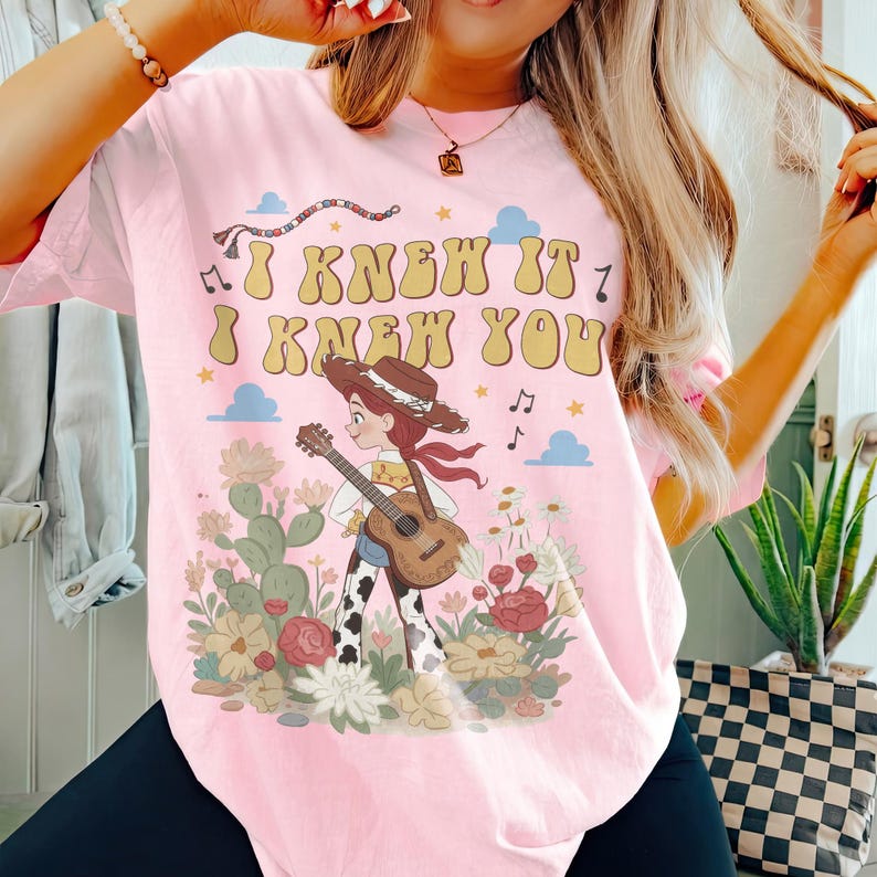 May include: A light pink t-shirt featuring a graphic of a cowgirl with a brown hat and red scarf playing an acoustic guitar. The text I KNEW IT I KNEW YOU is printed in yellow above the character, surrounded by cacti, flowers, clouds, and music notes.