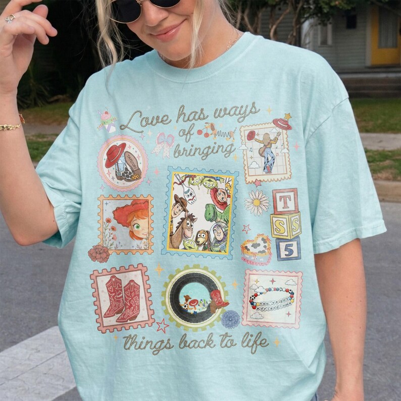 May include: A light blue t-shirt featuring a graphic print with the text "Love has ways of bringing things back to life" surrounded by various illustrations including characters from Toy Story, cowboy boots, alphabet blocks, and stars.