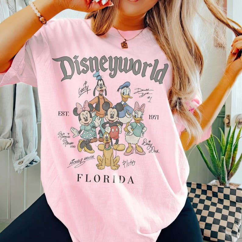 K&ouml;nnte beinhalten: Hellrosa T-Shirt mit der Aufschrift "Disneyworld Florida" und den Zeichentrickfiguren Mickey Mouse, Minnie Mouse, Goofy, Donald Duck, Daisy Duck und Pluto. Das Shirt hat auch den Text "EST. 1971".