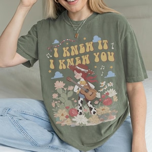 May include: An olive green short-sleeve t-shirt featuring a graphic print of a cowgirl character playing an acoustic guitar in a desert landscape with flowers and cacti. The text "I KNEW IT I KNEW YOU" is printed in a retro yellow font above.