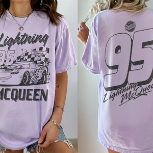 May include: Lavender t-shirt featuring a graphic of Lightning McQueen from the Cars movie. The front design includes the text "Lightning 95 McQueen" and a race car. The back displays a large "95" and "Lightning McQueen".
