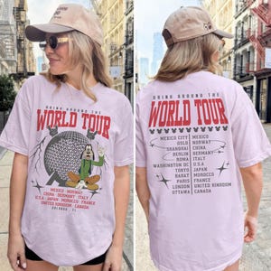May include: A light purple t-shirt with the text "DRINK AROUND THE WORLD TOUR" in red. The front features a graphic of a globe and a cartoon character. The back lists cities. The person is wearing a tan baseball cap.