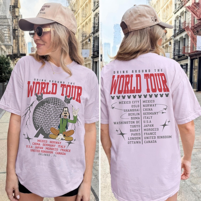 May include: A light purple t-shirt with the text "DRINK AROUND THE WORLD TOUR" in red. The front features a graphic of a globe and a cartoon character. The back lists cities. The person is wearing a tan baseball cap.