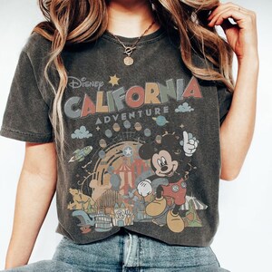 Puede incluir: Camiseta gris oscuro con el texto "Disney California Adventure" sobre un gr&aacute;fico colorido de Mickey Mouse, un cohete y una noria. La camiseta tiene un aspecto vintage y desgastado.