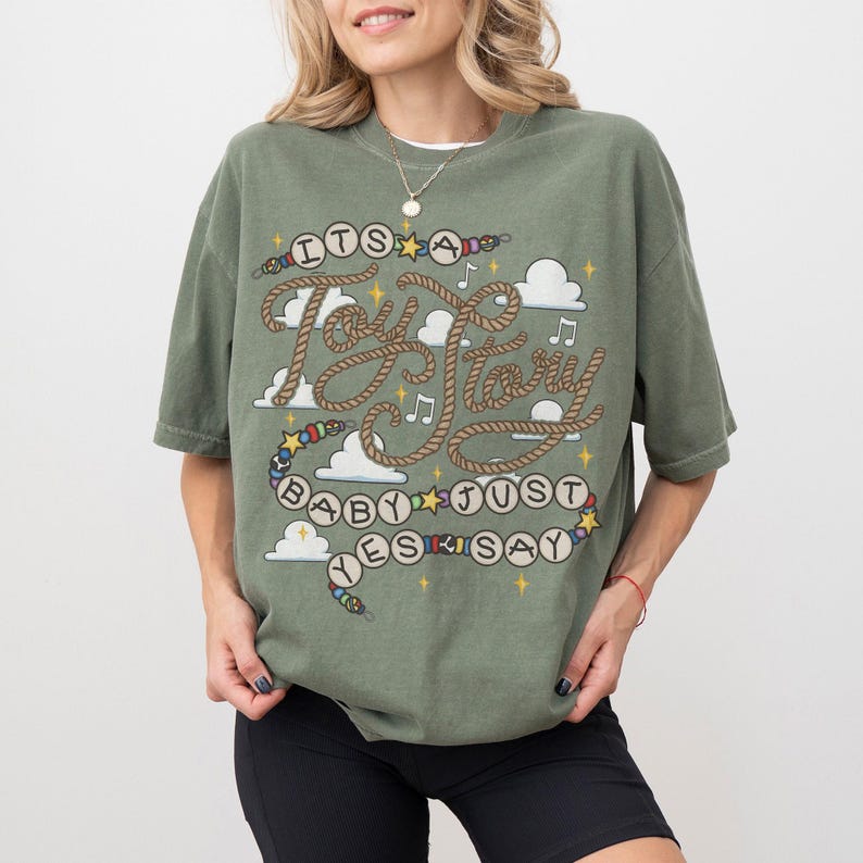 May include: An olive green short-sleeve t-shirt featuring a graphic design with the text "ITS A Toy Story BABY JUST SAY YES". The design includes rope-style lettering, colorful beads, white clouds, musical notes, and yellow stars.