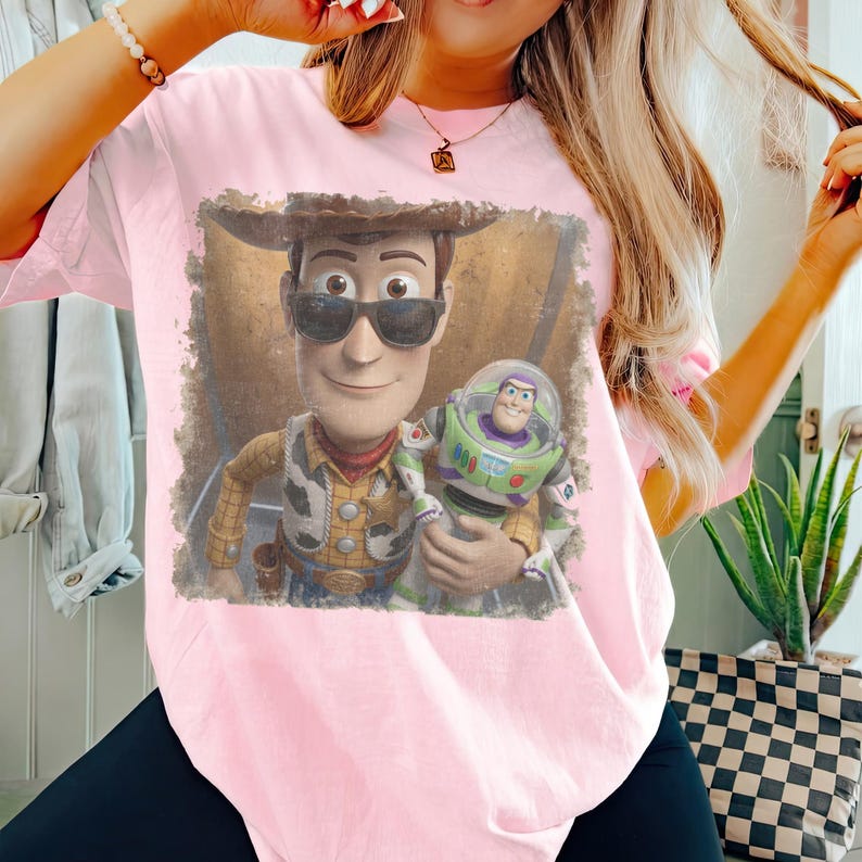 May include: A light pink short-sleeve t-shirt featuring a graphic print of the characters Woody and Buzz Lightyear. Woody wears black sunglasses and a cowboy hat, while Buzz Lightyear is held in the foreground. The graphic has a distressed border.