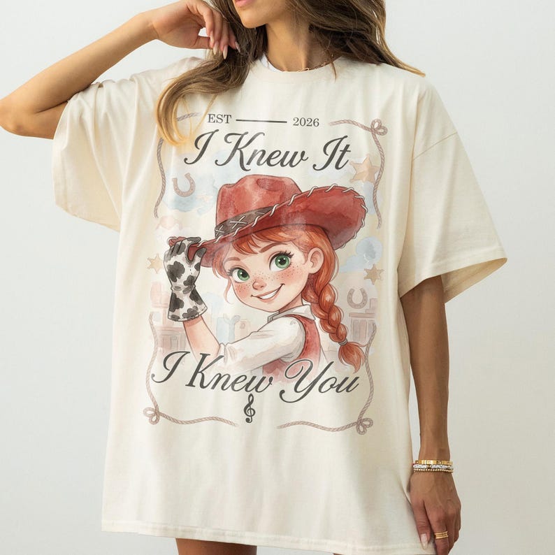 May include: An off-white short-sleeve t-shirt featuring a graphic of a person in a red cowboy hat and cow-print glove. The text reads "EST 2026", "I Knew It", and "I Knew You" with a rope border, stars, and horseshoe accents in a watercolor style.