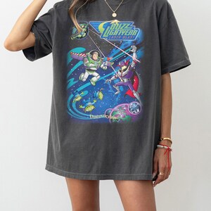 Puede incluir: Camiseta gris oscuro con un gr&aacute;fico colorido de Buzz Lightyear y Zurg de la pel&iacute;cula Toy Story. El dise&ntilde;o incluye el texto "Buzz Lightyear Laser Blast" y "Discoveryland".