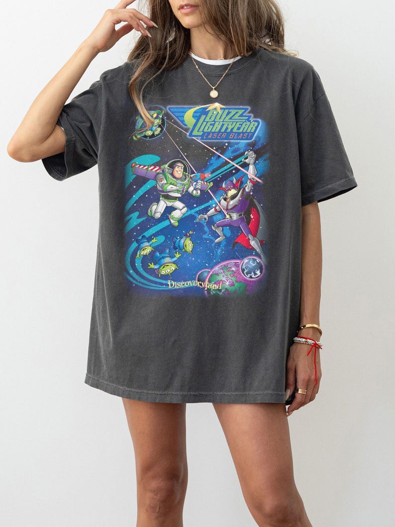 Puede incluir: Camiseta gris oscuro con un gr&aacute;fico colorido de Buzz Lightyear y Zurg de la pel&iacute;cula Toy Story. El dise&ntilde;o incluye el texto "Buzz Lightyear Laser Blast" y "Discoveryland".