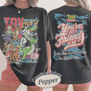 K&ouml;nnte beinhalten: Ein schwarzes Kurzarm-T-Shirt mit Toy Story-Grafiken. Die Vorderseite zeigt Jessie, Woody, Buzz, Slinky Dog, Bullseye und Hamm mit Text. Die R&uuml;ckseite zeigt das Toy Story-Logo, den Text You've Got A Friend In Me und die Namen der Figuren.