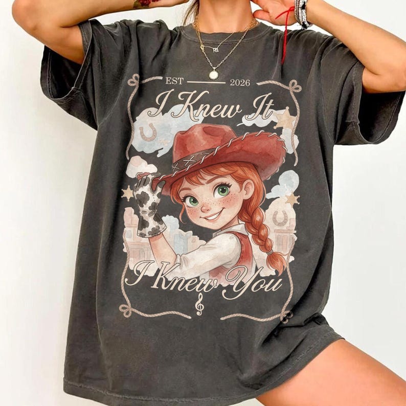 May include: A charcoal gray short-sleeve t-shirt featuring a graphic of a cartoon cowgirl with red hair and a brown hat. The text reads EST 2026, I Knew It, and I Knew You, framed by a rope design with stars, horseshoes, and a musical note.