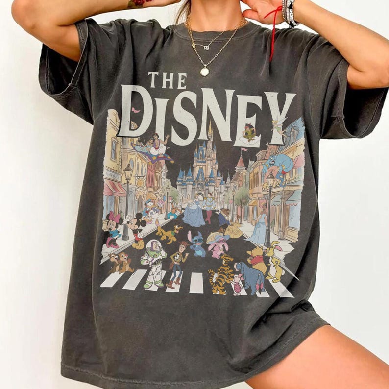 May include: Dark grey t-shirt with the words "THE DISNEY" and a colourful illustration of Disney characters crossing a street. The graphic includes Mickey Mouse, Buzz Lightyear, and Cinderella.