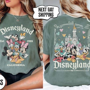May include: Moss green t-shirt featuring the Disneyland logo with Mickey Mouse, Minnie Mouse, Donald Duck, Goofy, and Daisy Duck. The front of the shirt has the Disneyland logo and characters. The back of the shirt has a castle and more characters.