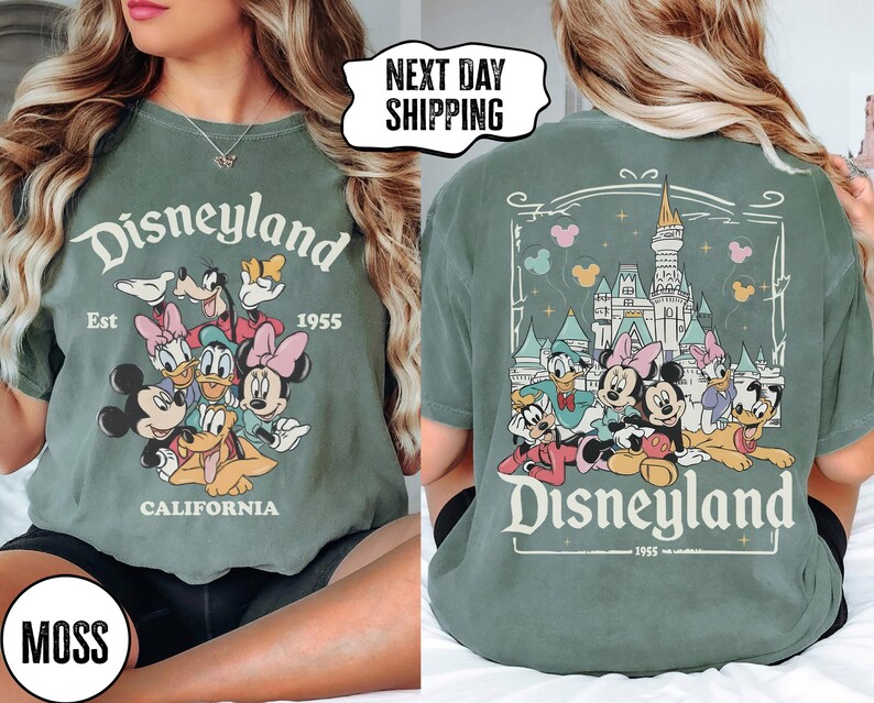 May include: Moss green t-shirt featuring the Disneyland logo with Mickey Mouse, Minnie Mouse, Donald Duck, Goofy, and Daisy Duck. The front of the shirt has the Disneyland logo and characters. The back of the shirt has a castle and more characters.