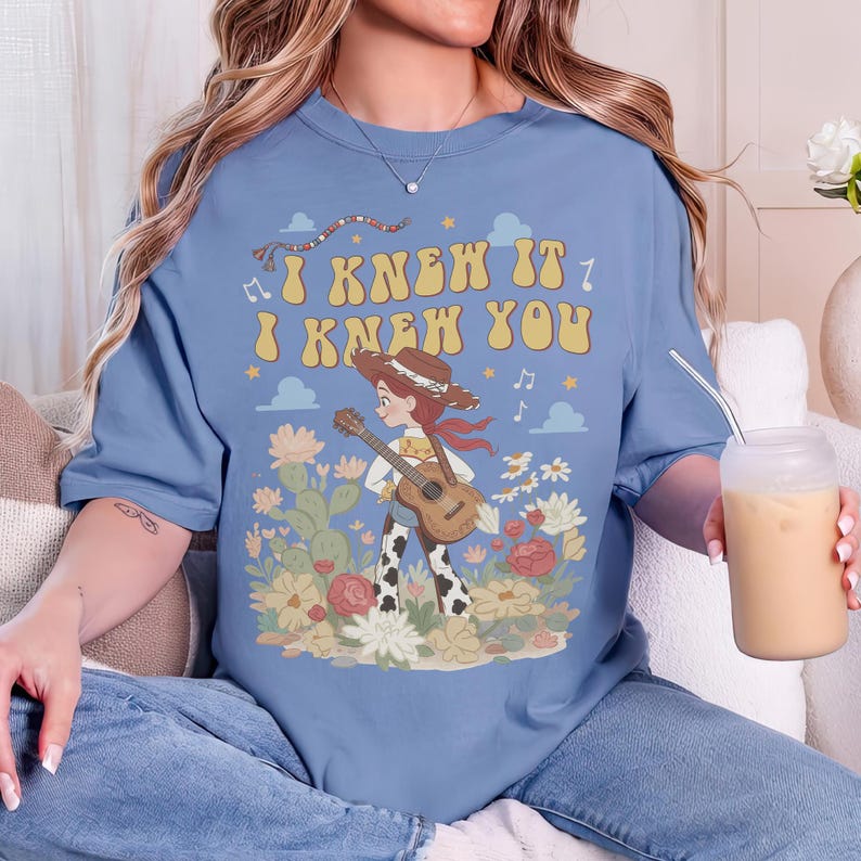 May include: A dusty blue short-sleeve t-shirt featuring a graphic print of a cowgirl playing an acoustic guitar in a desert landscape with cacti and flowers. The text "I KNEW IT I KNEW YOU" is printed in a retro yellow font above the illustration.