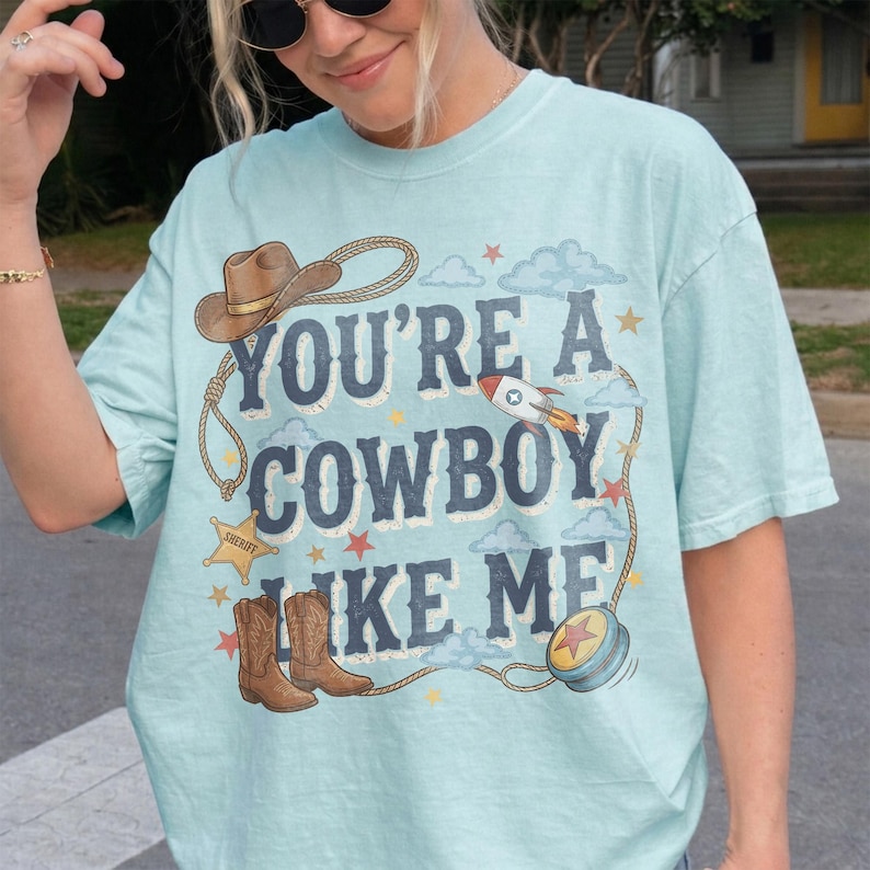 Puede incluir: Una camiseta de manga corta de color azul claro con un estampado gr&aacute;fico que incluye el texto "YOU'RE A COWBOY LIKE ME" en letras azul oscuro. El dise&ntilde;o incluye un sombrero de vaquero marr&oacute;n, una estrella de sheriff, botas y un cohete.