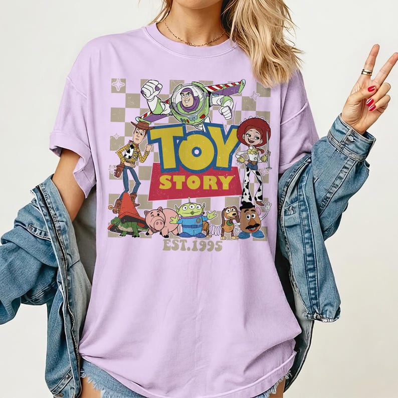 May include: A light purple t-shirt featuring the Toy Story logo and characters. The design includes Buzz Lightyear, Woody, Jessie, and other characters from the movie, with "EST.1995" printed below. The shirt has a vintage, distressed look.