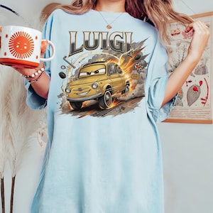 以下が含まれることがあります： 「LUIGI」の文字と、黄色の車の漫画イラストが描かれた水色のTシャツ。車はスピードラインと煙のエフェクトで動きが表現されています。