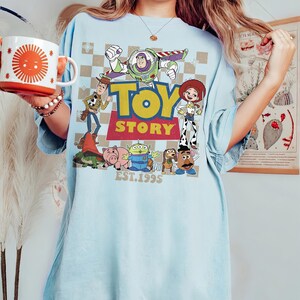 May include: Light blue t-shirt featuring a colorful graphic of Toy Story characters, including Buzz Lightyear, Woody, and Jessie. The words "TOY STORY" are prominently displayed, with "EST. 1995" below. A white and orange mug with a sun design is also visible.