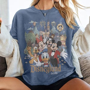 May include: Blue t-shirt featuring a Disneyland graphic with Mickey Mouse, Minnie Mouse, Donald Duck, Goofy, and other characters. The graphic includes the text "Disneyland Est. 1955" and a colorful illustration of the park.