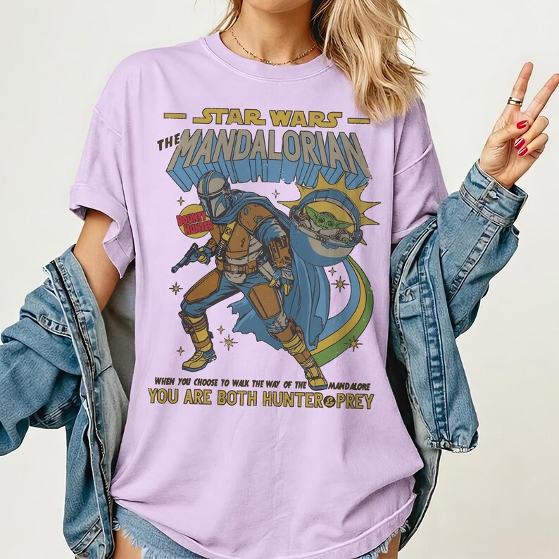 May include: A light purple t-shirt featuring a graphic of The Mandalorian from Star Wars. The design includes the text "STAR WARS THE MANDALORIAN" and the quote "WHEN YOU CHOOSE TO WALK THE WAY OF THE MANDALORE YOU ARE BOTH HUNTER PREY."