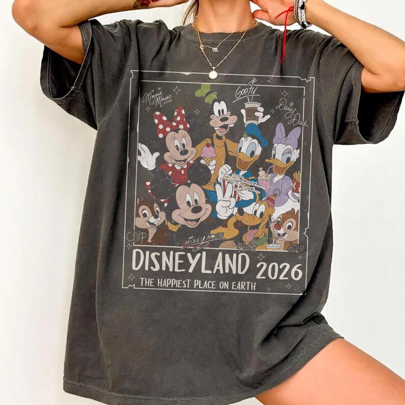 May include: Dark gray t-shirt featuring a Disneyland 2026 graphic with various Disney characters. The graphic includes Mickey Mouse, Minnie Mouse, Goofy, Donald Duck, Daisy Duck, Chip, and Dale. The text reads "Disneyland 2026" and "The Happiest Place on Earth."