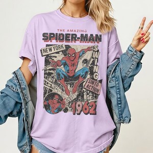 Op de afbeelding: Lichtpaars T-shirt met een retro Spider-Man-afbeelding. Het ontwerp bevat de tekst "THE AMAZING SPIDER-MAN", "NEW YORK" en "1962". Het shirt heeft korte mouwen en een losse pasvorm, ideaal voor casual wear.