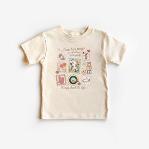 May include: A cream-colored short-sleeve t-shirt featuring a central graphic print of various small illustrations including cowboy boots, a record, and building blocks, with the text "Love has ways of bringing things back to life" in brown.