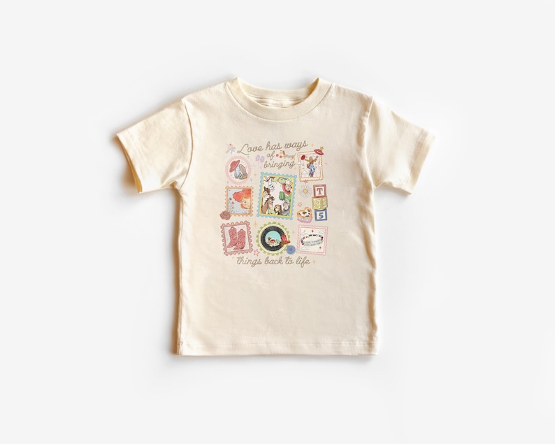 May include: A cream-colored short-sleeve t-shirt featuring a central graphic print of various small illustrations including cowboy boots, a record, and building blocks, with the text "Love has ways of bringing things back to life" in brown.