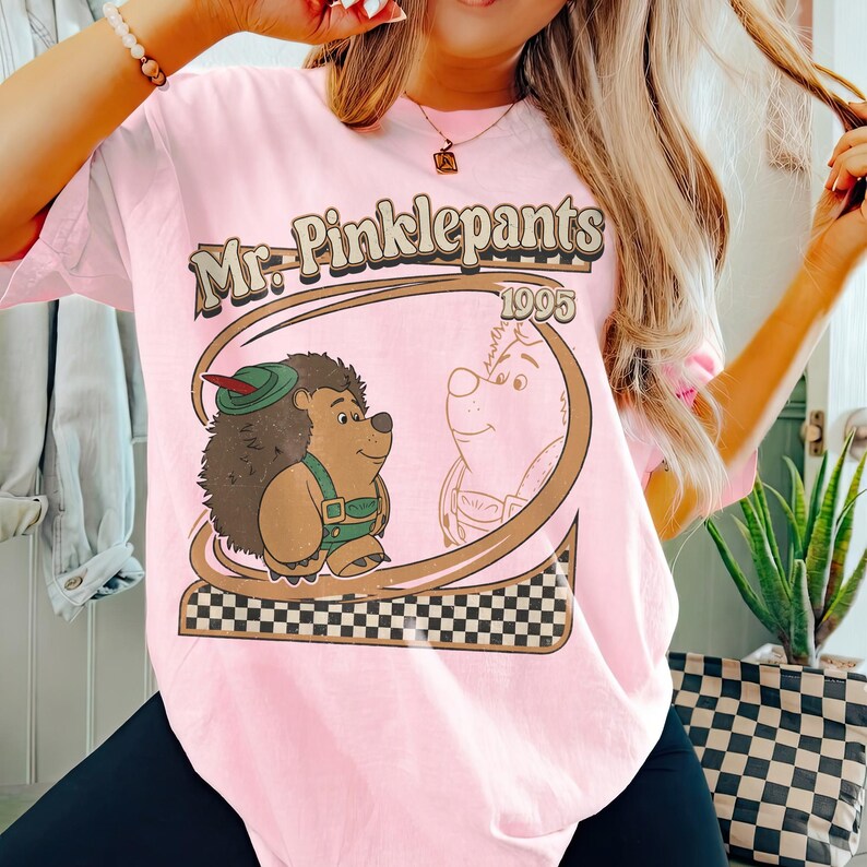 Puede incluir: Camiseta rosa claro con un dise&ntilde;o de dibujos animados que dice "Mr. Pinklepants 1995". El dise&ntilde;o incluye un erizo con sombrero verde y peto, y un oso. El dise&ntilde;o est&aacute; enmarcado por un &oacute;valo marr&oacute;n con un patr&oacute;n de tablero de ajedrez.