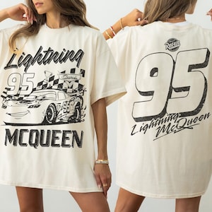 May include: Cream-colored t-shirt featuring a black graphic of Lightning McQueen race car with the number 95 and the name Lightning McQueen. The back of the shirt displays a large number 95 and the words Lightning McQueen.