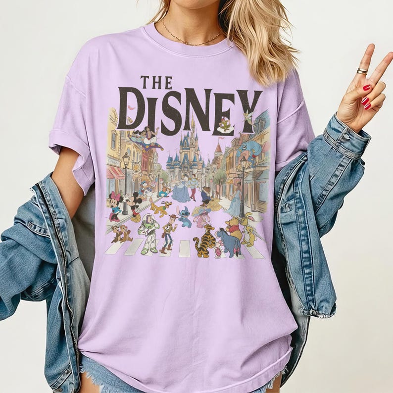 May include: A lilac t-shirt with the words "THE DISNEY" printed across the top. Below is a colourful illustration of Disney characters, including Mickey Mouse, Buzz Lightyear, and Winnie the Pooh, crossing a street.