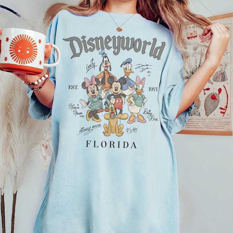 K&ouml;nnte beinhalten: Hellblaues T-Shirt mit einem Disneyworld-Motiv mit Micky Maus, Minnie Maus, Donald Duck, Daisy Duck, Goofy und Pluto. Die Grafik enth&auml;lt den Text "Disneyworld Florida" und "Est. 1971".