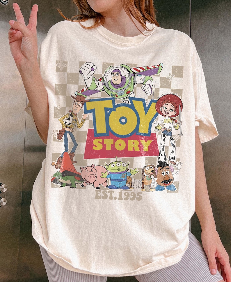 May include: Cream-colored t-shirt featuring a colorful graphic of characters from the animated film "Toy Story." The design includes Buzz Lightyear, Woody, Jessie, and other characters, with the words "TOY STORY" and "EST. 1995" printed on the front.