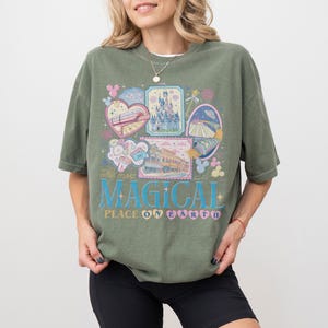 May include: Olive green t-shirt with a colorful graphic design. The design includes illustrations of a castle, a train, and other theme park elements. Text on the shirt reads "The most MAGICAL PLACE ON EARTH."