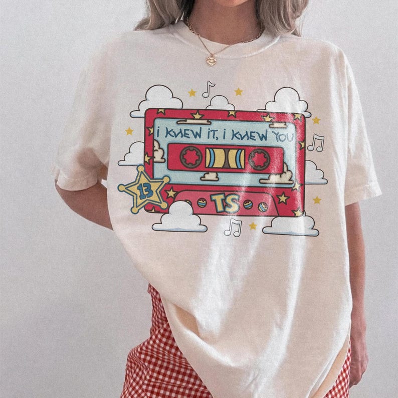 May include: A cream-colored short-sleeve t-shirt featuring a graphic of a red cassette tape surrounded by white clouds, yellow stars, and musical notes. The tape displays the text "I KNEW IT, I KNEW YOU" and "TS" with a star icon labeled "13".