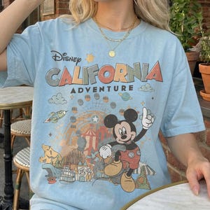 Puede incluir: Camiseta azul claro con el texto "Disney CALIFORNIA ADVENTURE" y un dibujo de Mickey Mouse y un parque tem&aacute;tico. La camiseta tiene un aspecto vintage descolorido.