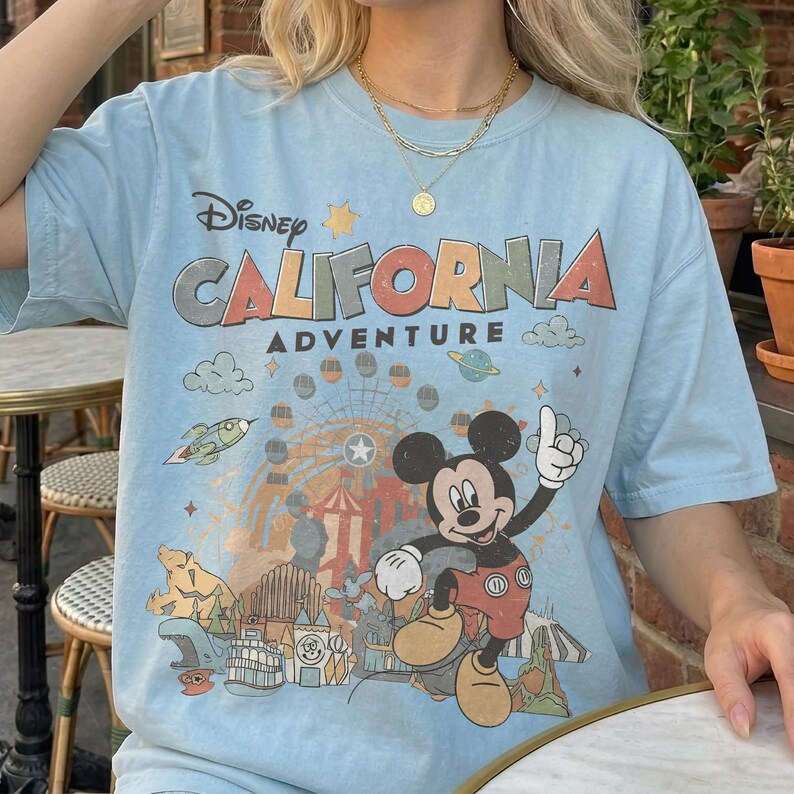 Puede incluir: Camiseta azul claro con el texto "Disney CALIFORNIA ADVENTURE" y un dibujo de Mickey Mouse y un parque tem&aacute;tico. La camiseta tiene un aspecto vintage descolorido.