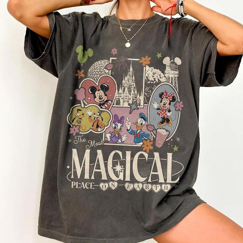 May include: Dark gray t-shirt featuring a colorful graphic of Disney characters and landmarks. The design includes Mickey and Minnie Mouse, Donald and Daisy Duck, and the text "The Most Magical Place on Earth."